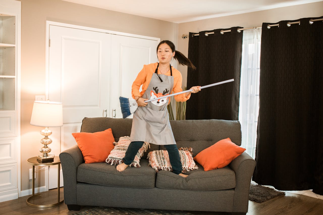 brand-01 A joyful woman dances with a broom while cleaning her cozy living room.